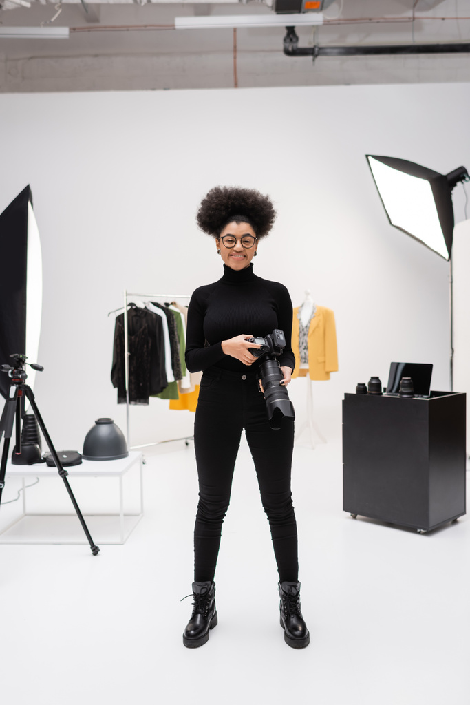 Modern fotoğraf stüdyosunda elinde dijital kamerayla dikilen siyah elbiseli Afro-Amerikan içerikli tam boy. - Fotoğraf, Görsel