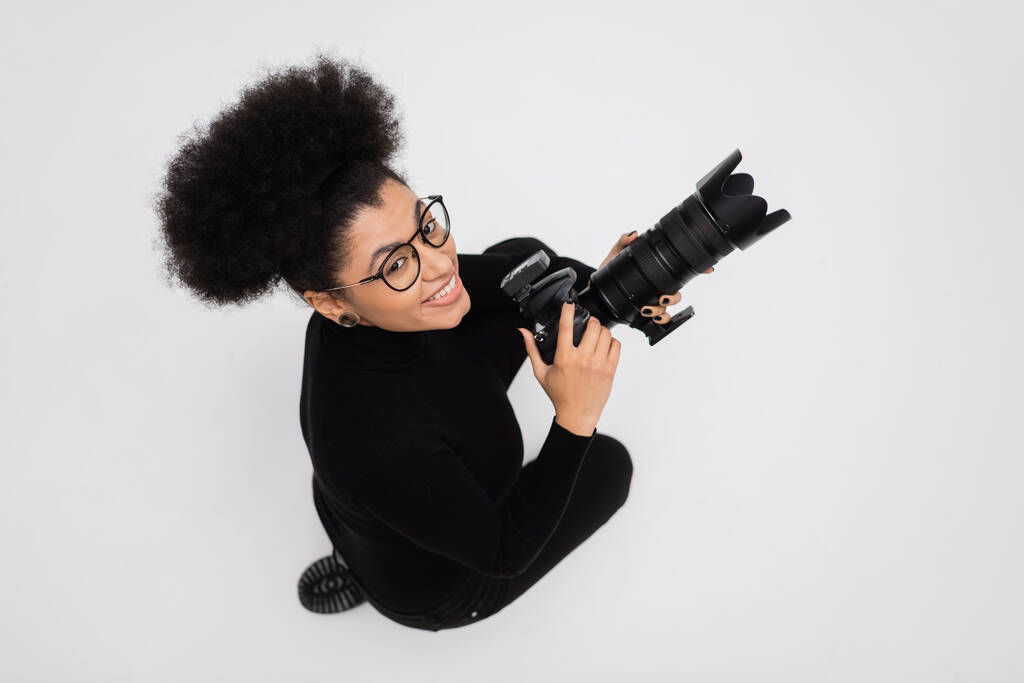 κορυφαία άποψη του υπερευχαριστημένου αφροαμερικανού παραγωγού περιεχομένου σε γυαλιά ηλίου που κατέχει επαγγελματική ψηφιακή φωτογραφική μηχανή απομονωμένη σε γκρι - Φωτογραφία, εικόνα