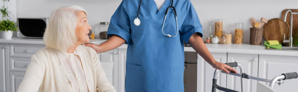 infirmière multiraciale en uniforme bleu avec stéthoscope debout près de la femme âgée et marchette, bannière  - Photo, image
