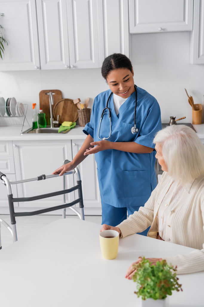 gelukkig multiraciale verpleegster in blauw uniform wijzend met de hand op walker terwijl staan in de buurt van senior vrouw met grijs haar  - Foto, afbeelding
