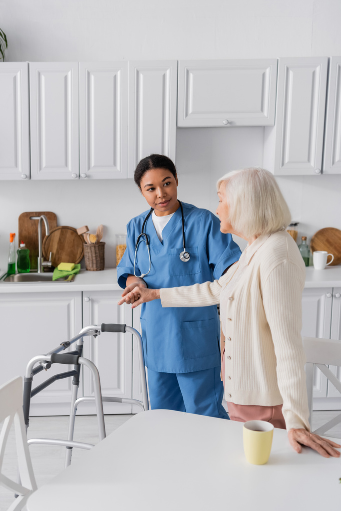 brünette, multiethnische Pflegerin in blauer Uniform unterstützt Seniorin mit grauen Haaren, während sie neben Rollator steht  - Foto, Bild