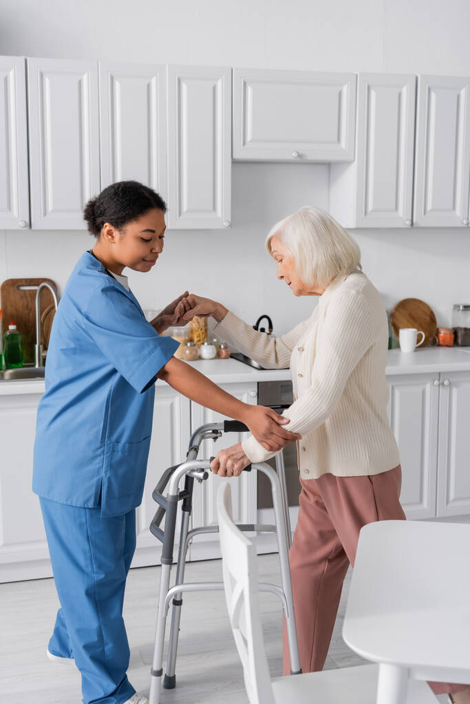 brunette multiracial nurse in blue uniform supporting senior woman with grey hair standing near walker  - Photo, Image