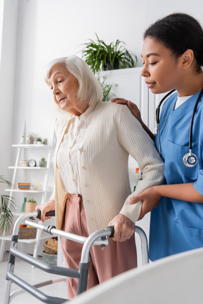fürsorgliche multiethnische Krankenschwester in blauer Uniform unterstützt eine Seniorin mit grauen Haaren beim Gehen mit Hilfe eines Rollators zu Hause  - Foto, Bild