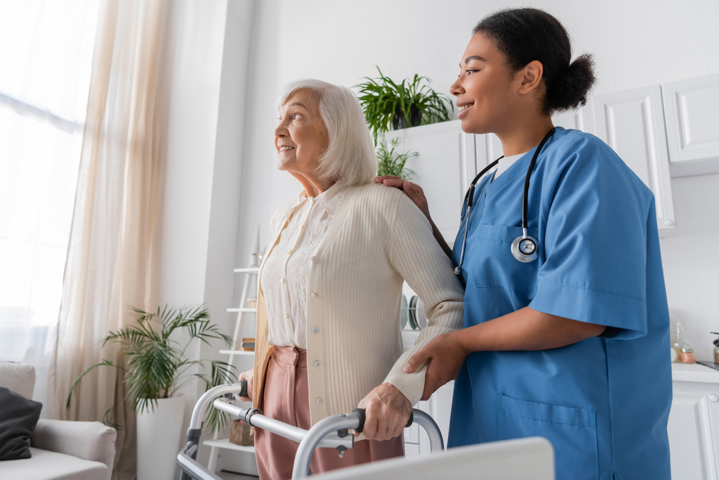 enfermera multirracial alegre en uniforme azul que apoya a mujer mayor feliz con el pelo gris que camina con la ayuda del caminante en el país  - Foto, imagen