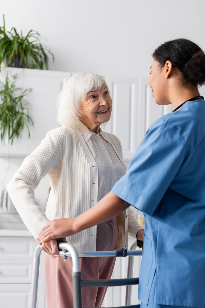 mulher sênior feliz com cabelos grisalhos andando com quadro walker e olhando para enfermeira multirracial morena  - Foto, Imagem