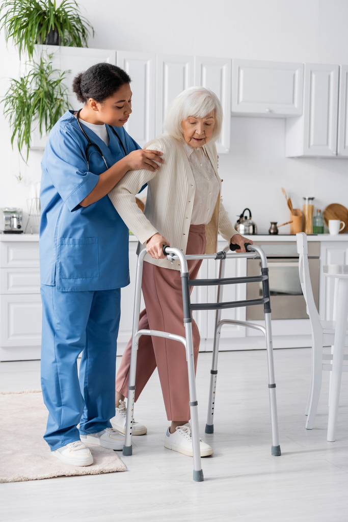 повна довжина багаторасової медсестри в уніформі підтримуючої пенсіонерки з сірим волоссям, що йде з прогулянковою рамою вдома
  - Фото, зображення