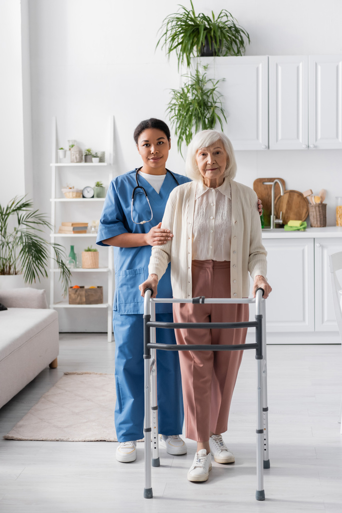 引退した女性をサポートする多人種看護師の長さ灰色の髪の家で歩くフレームと歩く  - 写真・画像