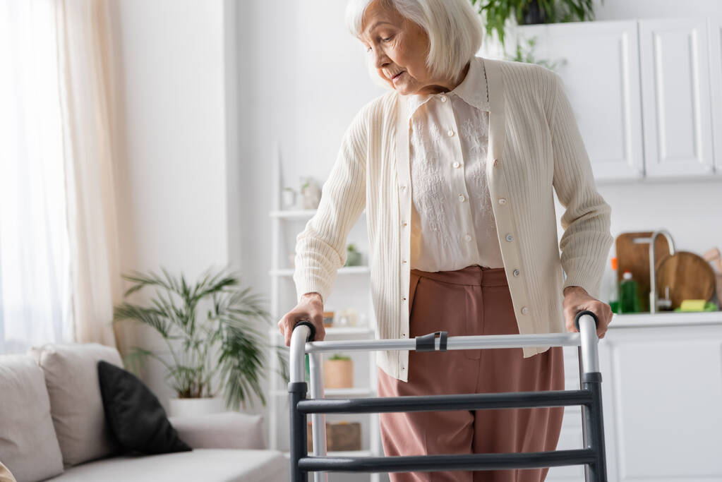 ηλικιωμένη γυναίκα με γκρίζα μαλλιά περπάτημα με τη βοήθεια του περιπατητή στο σύγχρονο διαμέρισμα  - Φωτογραφία, εικόνα