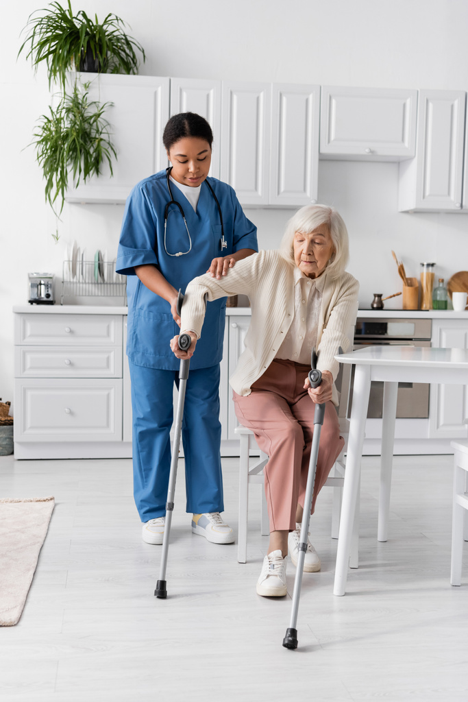 πλήρες μήκος της πολύφυλης νοσοκόμας με στολή βοηθώντας ηλικιωμένη γυναίκα χρησιμοποιώντας πατερίτσες να σηκωθεί  - Φωτογραφία, εικόνα