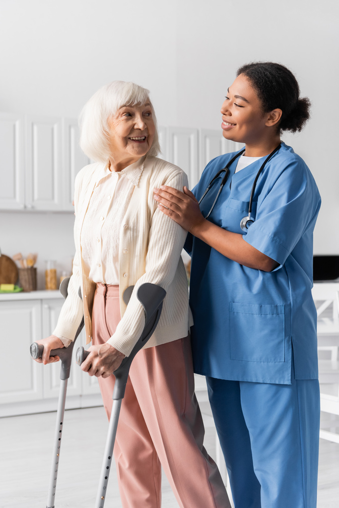 χαρούμενη συνταξιούχος γυναίκα με γκρίζα μαλλιά χρησιμοποιώντας πατερίτσες ενώ περπατούσε κοντά σε πολυφυλετική νοσοκόμα στο σπίτι  - Φωτογραφία, εικόνα