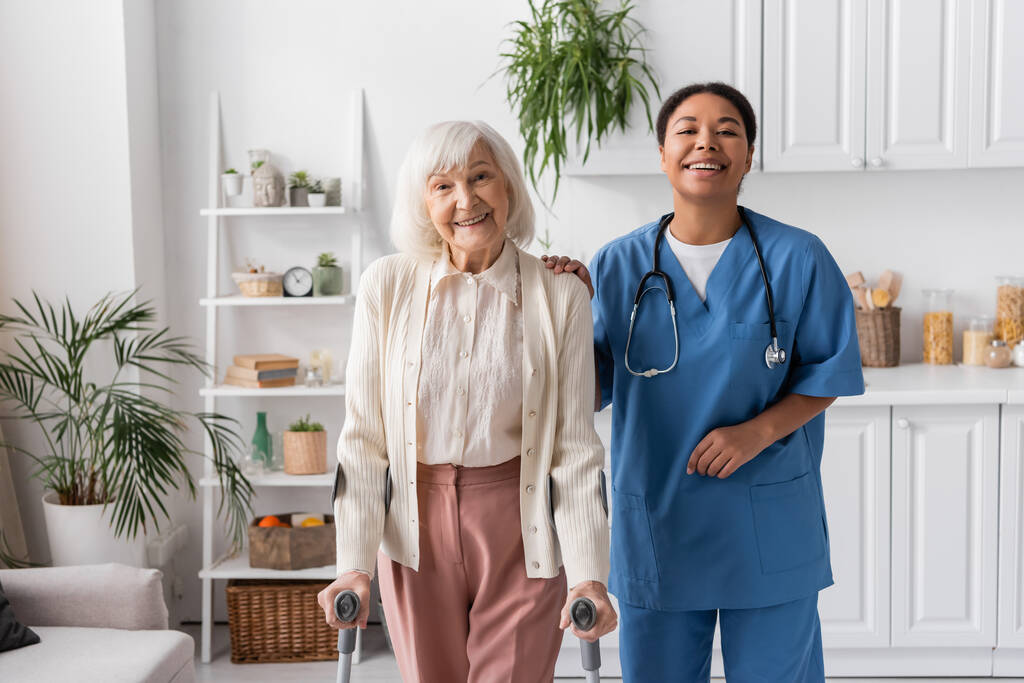 mujer jubilada positiva con pelo gris usando muletas mientras camina cerca de la enfermera multirracial feliz en casa  - Foto, imagen