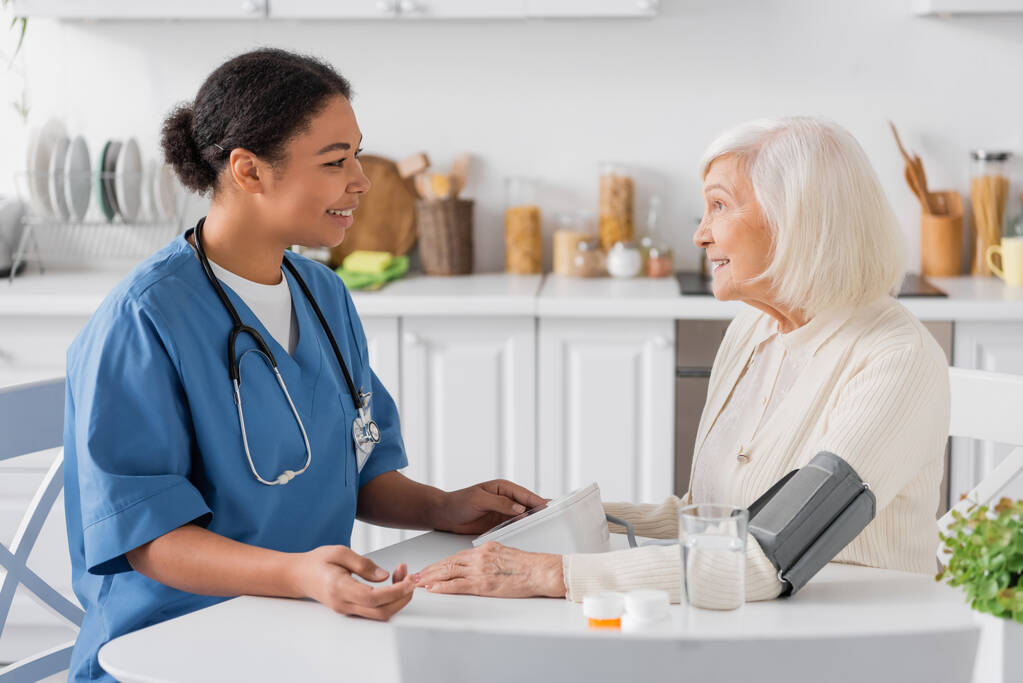 fröhliche multiethnische Krankenschwester misst den Blutdruck einer Seniorin mit grauen Haaren neben Medikamenten auf dem Tisch   - Foto, Bild