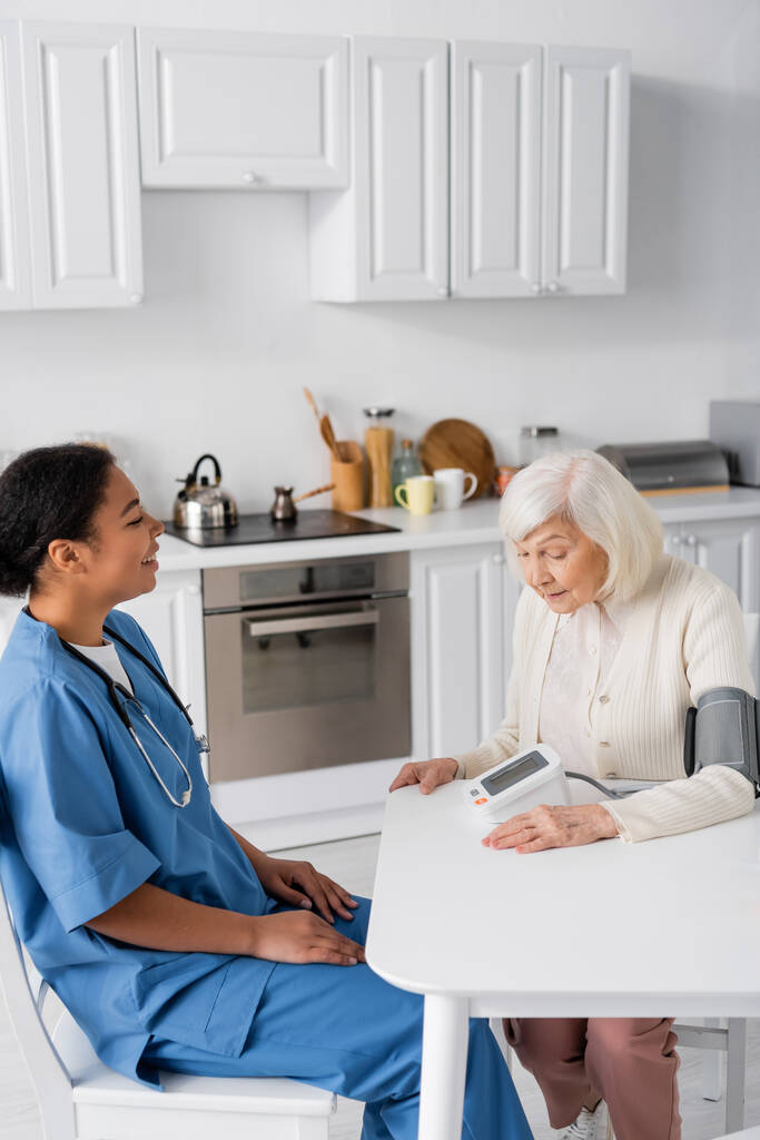 felice infermiera multirazziale guardando la donna anziana con i capelli grigi misurazione della pressione sanguigna  - Foto, immagini