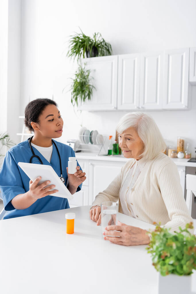 enfermeira multirracial morena usando tablet digital e segurando medicação enquanto conversa com uma mulher aposentada com cabelos grisalhos - Foto, Imagem