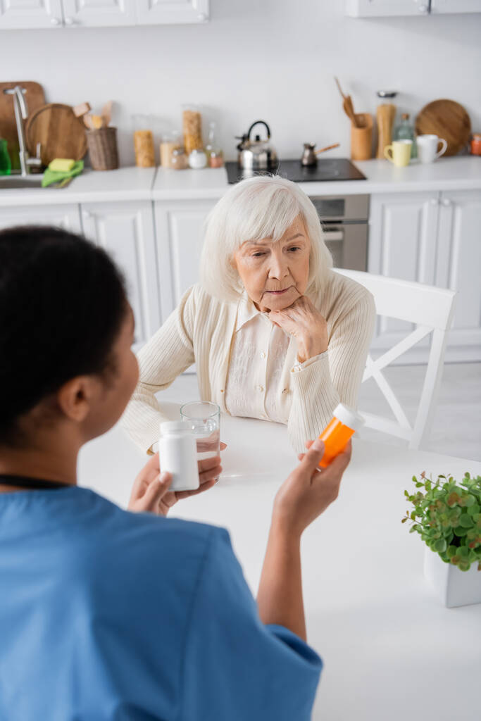vista aérea de la enfermera multirracial morena sosteniendo medicación mientras habla con una mujer jubilada con el pelo gris - Foto, Imagen