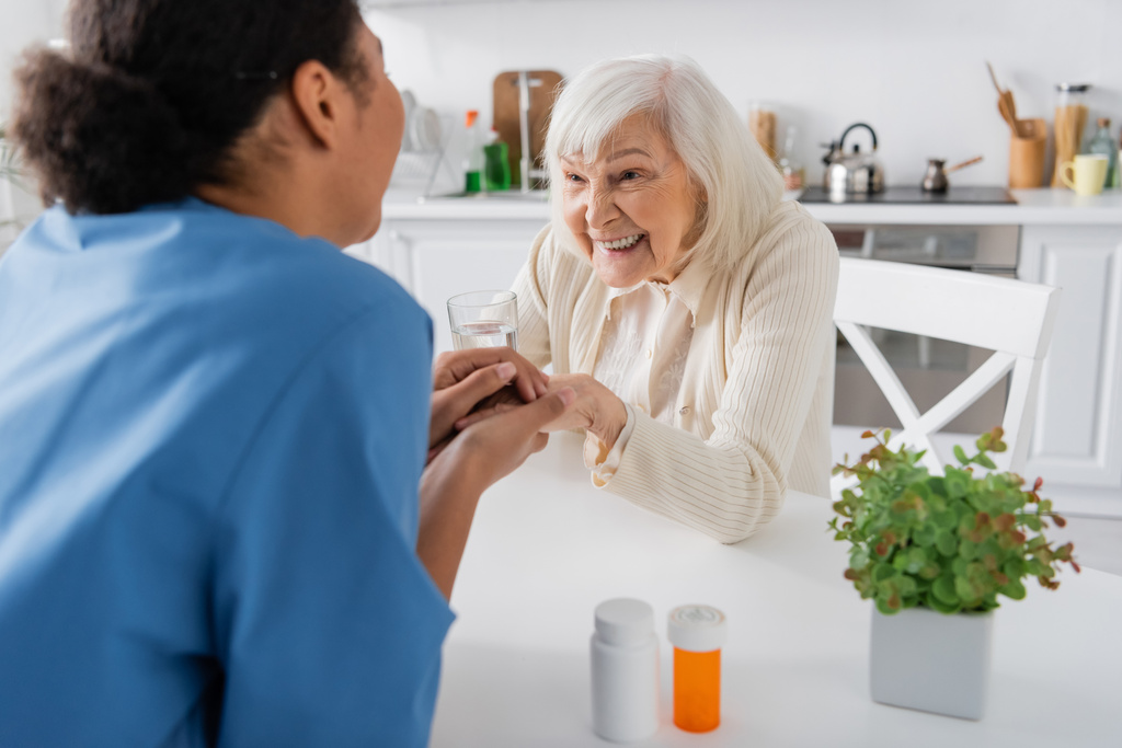 wielorasowa pielęgniarka trzymająca się za ręce ze szczęśliwą starszą kobietą obok leków na stole  - Zdjęcie, obraz