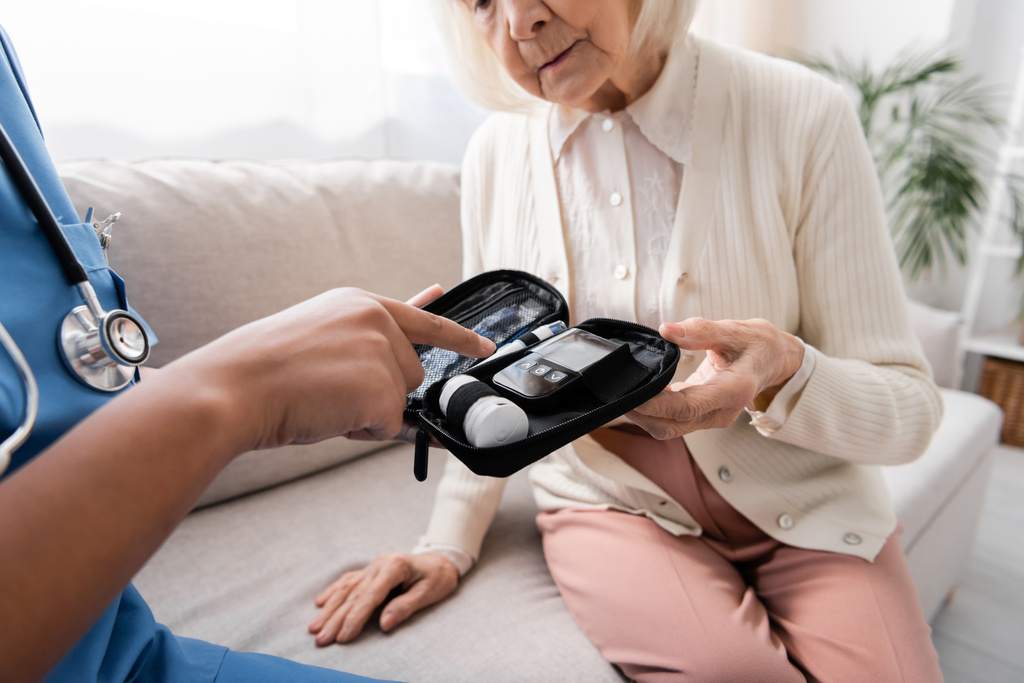 visão cortada de enfermeira multirracial em uniforme apontando para kit de diabetes perto de mulher idosa sentada no sofá  - Foto, Imagem