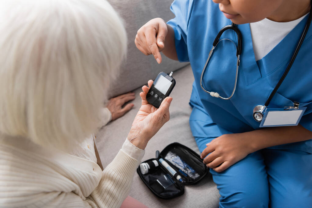 oříznutý pohled na multirakiální pečovatelku v uniformě ukazující na glukometr poblíž starší ženy s šedivými vlasy  - Fotografie, Obrázek