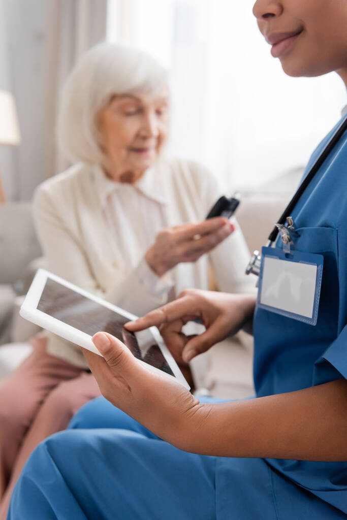 πολυφυλετική νοσοκόμα χρησιμοποιώντας ψηφιακό δισκίο με λευκή οθόνη κοντά στην ηλικιωμένη γυναίκα σε θολή φόντο  - Φωτογραφία, εικόνα