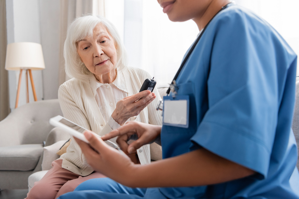 Seniorin mit grauen Haaren hält Glukometer in der Nähe einer Krankenschwester mit digitalem Tablet  - Foto, Bild