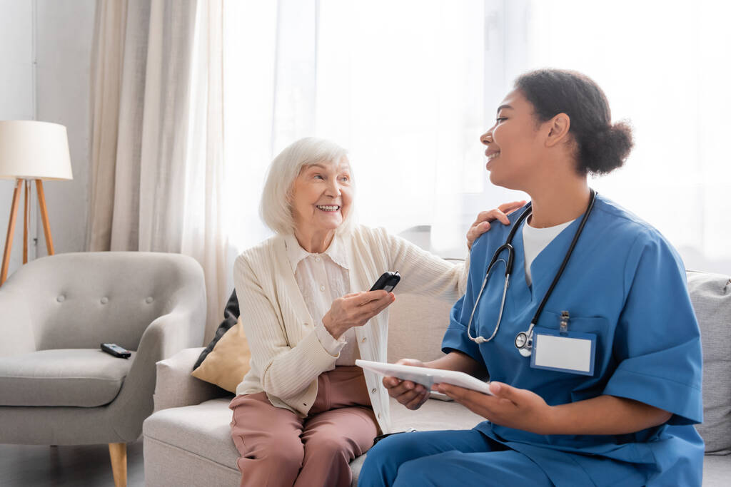 веселая пожилая женщина с седыми волосами, держащая глюкометр рядом с счастливой многорасовой медсестрой с помощью цифрового планшета  - Фото, изображение
