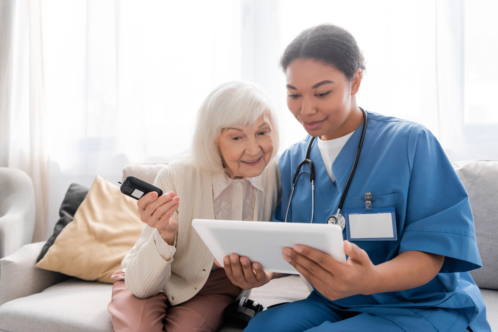 Χαρούμενη ηλικιωμένη γυναίκα με γκρίζα μαλλιά κρατώντας γλυκομετρικό και κοιτάζοντας ψηφιακή ταμπλέτα κοντά στην ευτυχισμένη πολυφυλετική νοσοκόμα  - Φωτογραφία, εικόνα