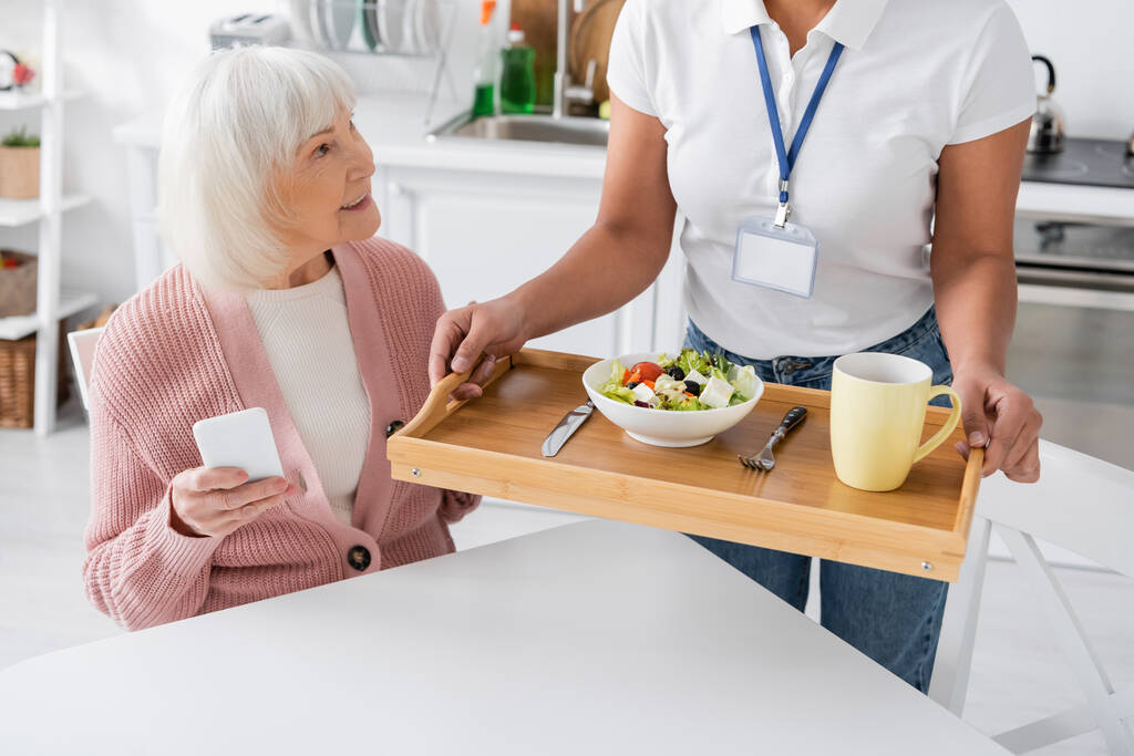 travailleur social multiracial tenant plateau avec déjeuner pour femme âgée heureuse avec les cheveux gris  - Photo, image