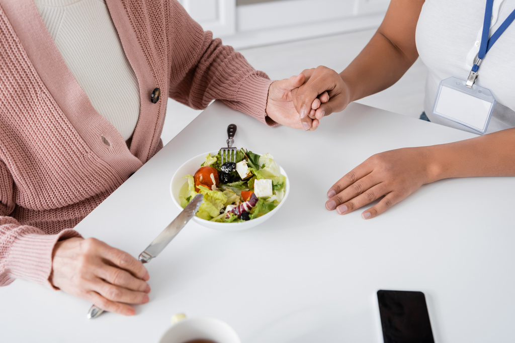 vue du dessus de la femme âgée tenant la main avec l'aidant multiracial à côté du déjeuner sur la table  - Photo, image