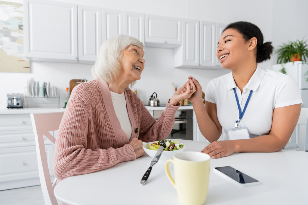 heureuse femme retraitée avec les cheveux gris tenant la main avec travailleur social multiracial joyeux à côté du déjeuner sur la table  - Photo, image