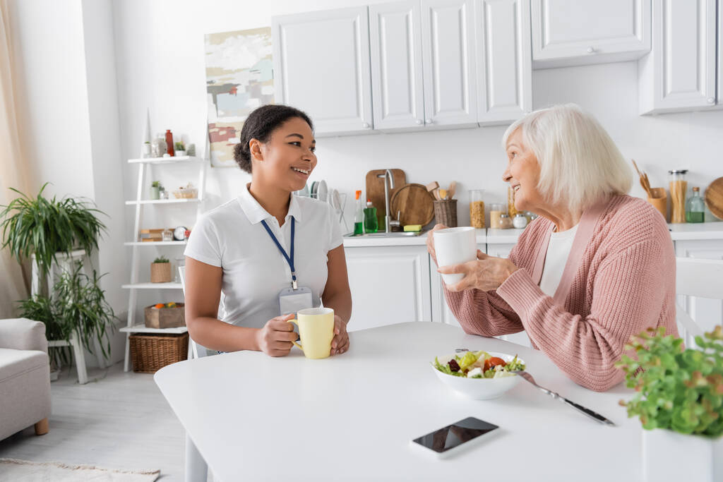 fröhliche multiethnische Sozialarbeiterin beim Tee mit Seniorin in der Küche  - Foto, Bild