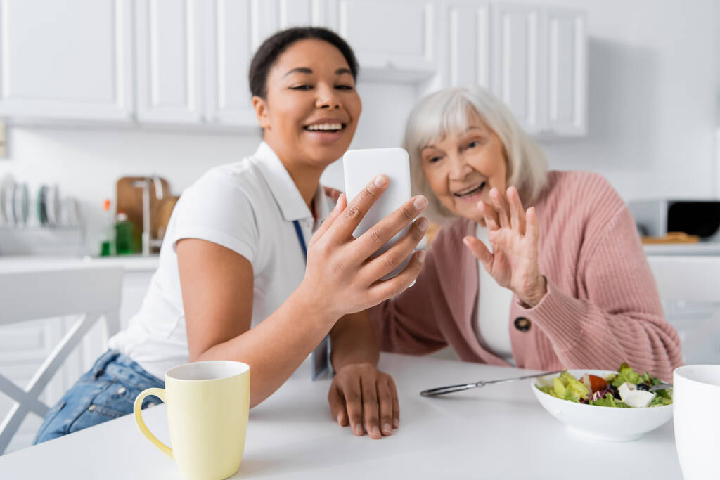 szczęśliwy wielorasowy pracownik socjalny gospodarstwa smartfon podczas senior kobieta macha ręką podczas rozmowy wideo  - Zdjęcie, obraz