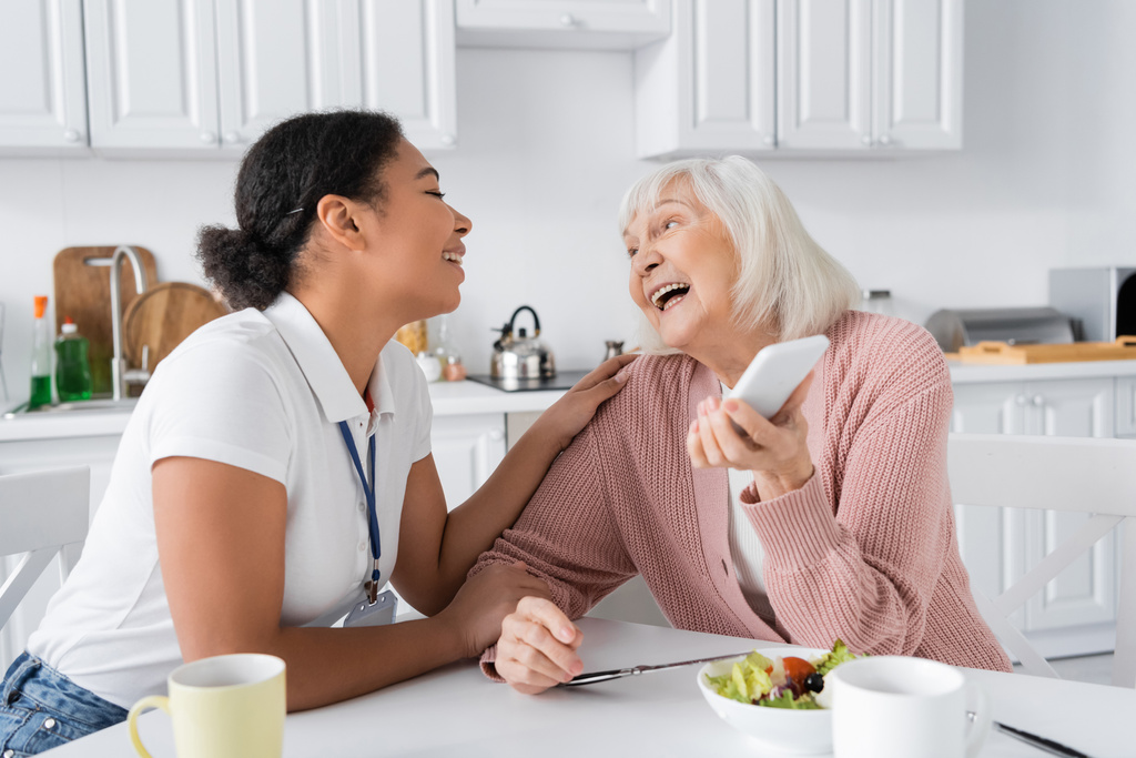 счастливая пожилая женщина со смартфоном рядом с веселым мультирасовым социальным работником дома  - Фото, изображение