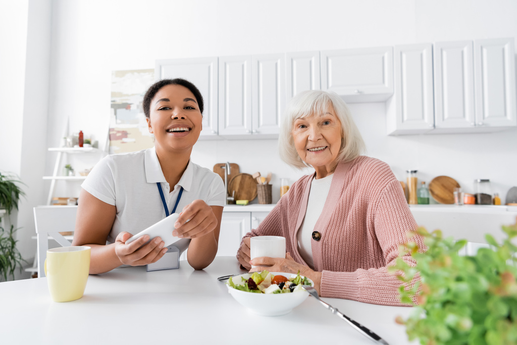 feliz trabajador social multirracial sosteniendo teléfono inteligente durante el almuerzo con la mujer jubilada en la cocina  - Foto, Imagen