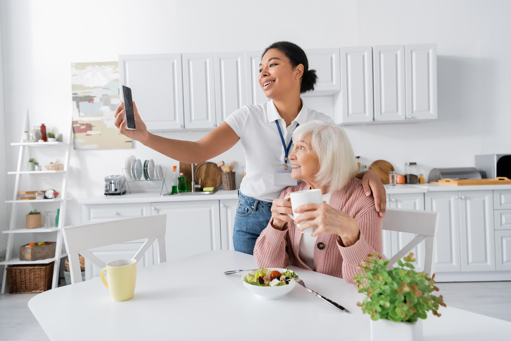 fröhliche multiethnische Sozialarbeiterin macht Selfie mit Rentnerin während des Mittagessens in der Küche  - Foto, Bild