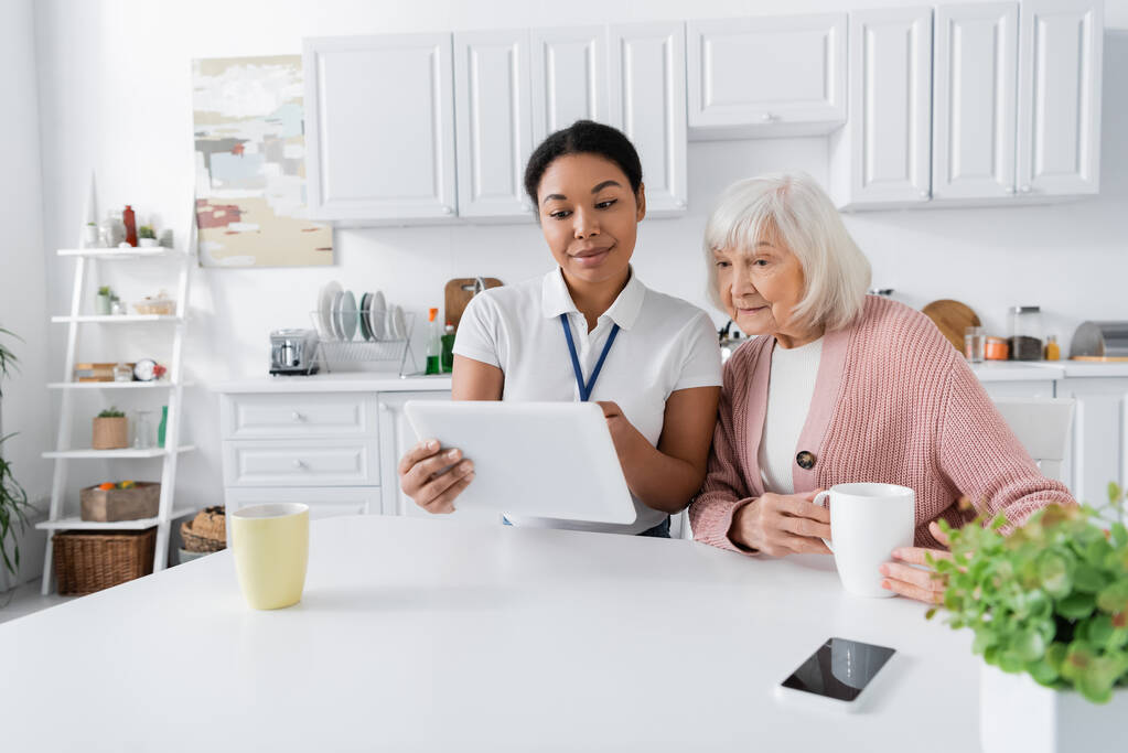 Glückliche Sozialarbeiterin hält digitales Tablet neben Seniorin in Küche  - Foto, Bild