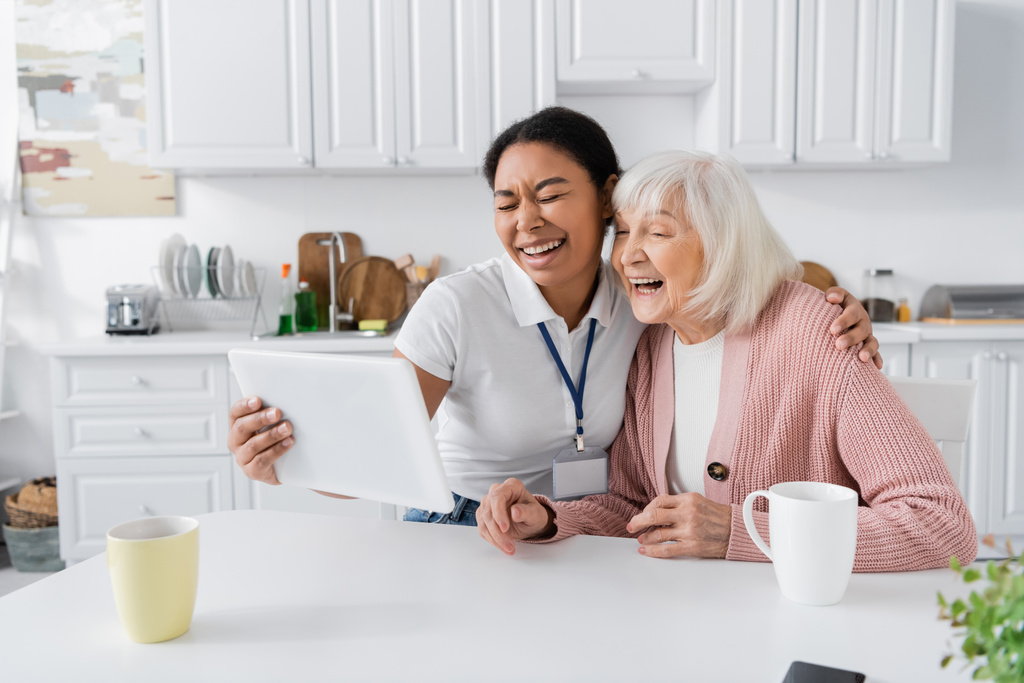 onnellinen monirotuinen sosiaalityöntekijä tilalla digitaalinen tabletti ja nauraa vanhempi nainen keittiössä  - Valokuva, kuva