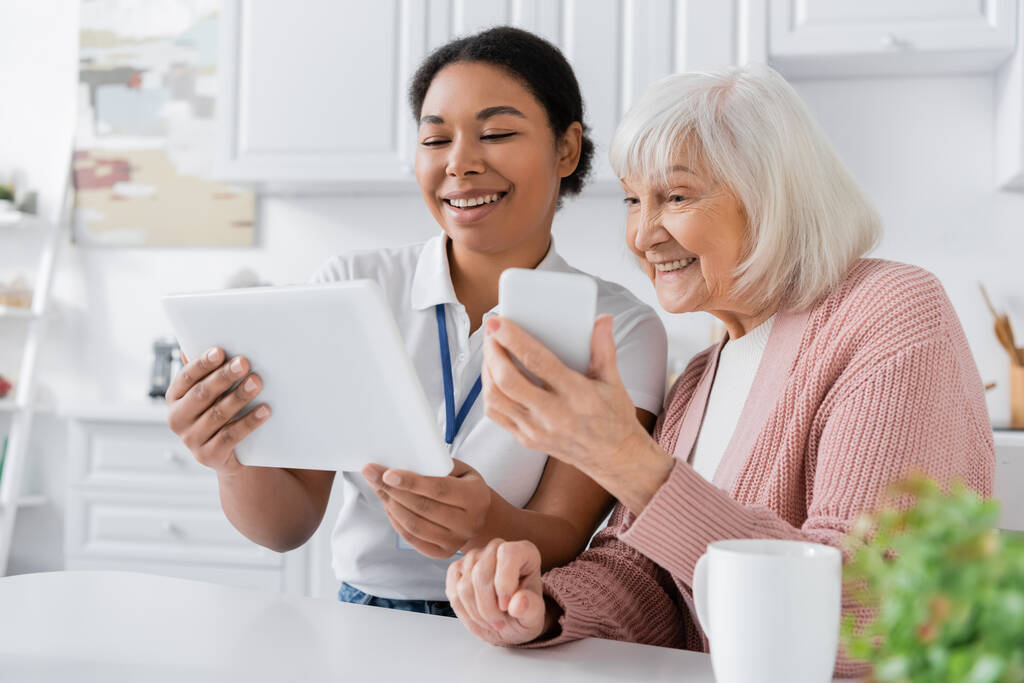 Mutfakta akıllı telefonu olan yaşlı bir kadının yanında dijital tablet tutan mutlu sosyal hizmet görevlisi.  - Fotoğraf, Görsel