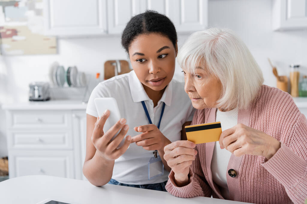 morena multirracial trabajador social sosteniendo teléfono inteligente cerca de la mujer mayor con tarjeta de crédito  - Foto, Imagen
