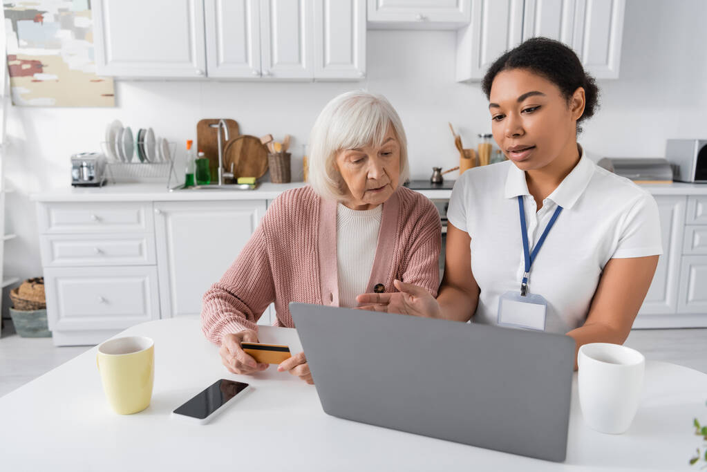 Sozialarbeiterin mit Laptop erklärt Seniorin mit Kreditkarte, wie man online einkauft  - Foto, Bild
