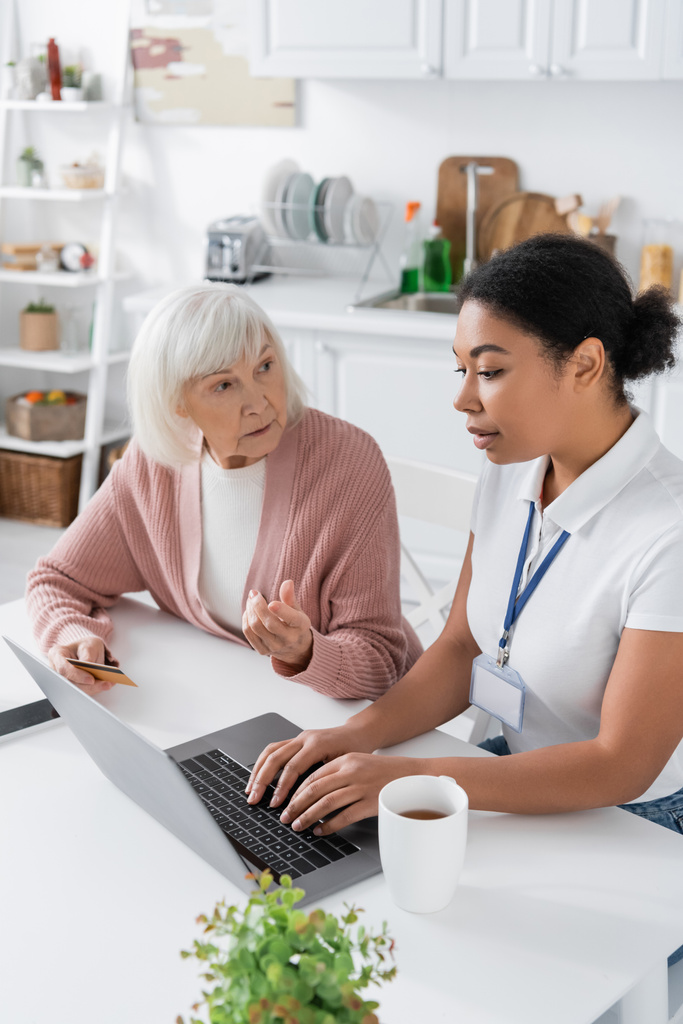 multiraciale maatschappelijk werker met behulp van laptop in de buurt van senior vrouw met creditcard  - Foto, afbeelding