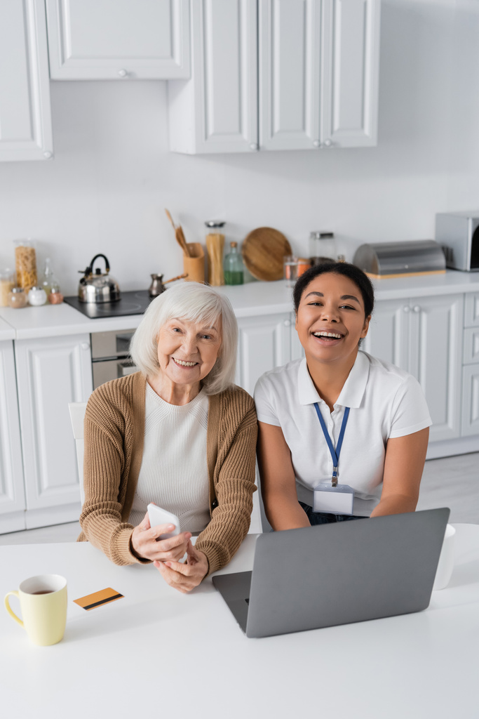 Fröhliche Sozialarbeiterin und Seniorin halten Smartphone neben Laptop auf Tisch  - Foto, Bild