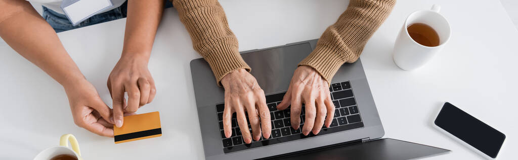 κορυφαία άποψη της τρίτης γυναίκας πληκτρολογώντας στο πληκτρολόγιο laptop κοντά πολυφυλετική κοινωνική λειτουργός με πιστωτική κάρτα, πανό  - Φωτογραφία, εικόνα