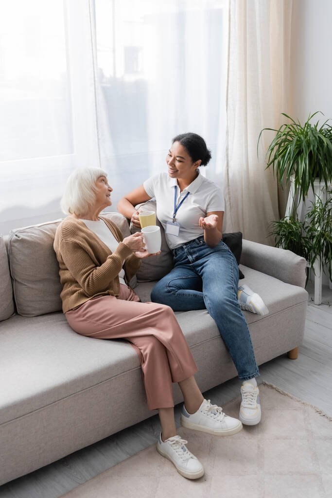 fröhliche multirassische Sozialarbeiterin plaudert mit Seniorin beim Tee im Wohnzimmer  - Foto, Bild