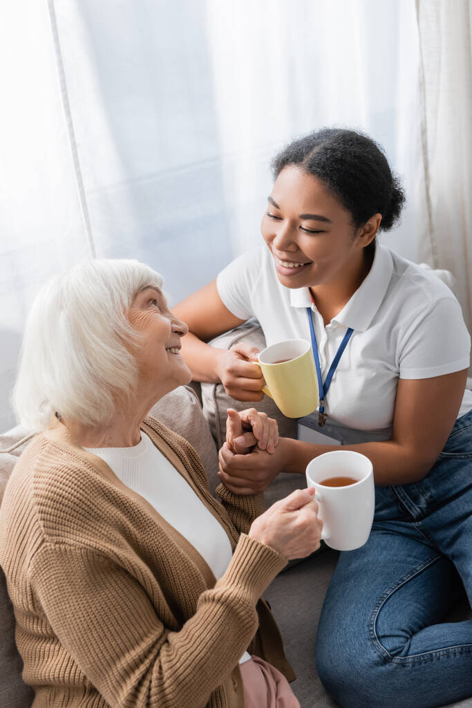 wesoły wielorasowy pracownik socjalny czatujący ze starszą kobietą podczas picia herbaty w salonie  - Zdjęcie, obraz