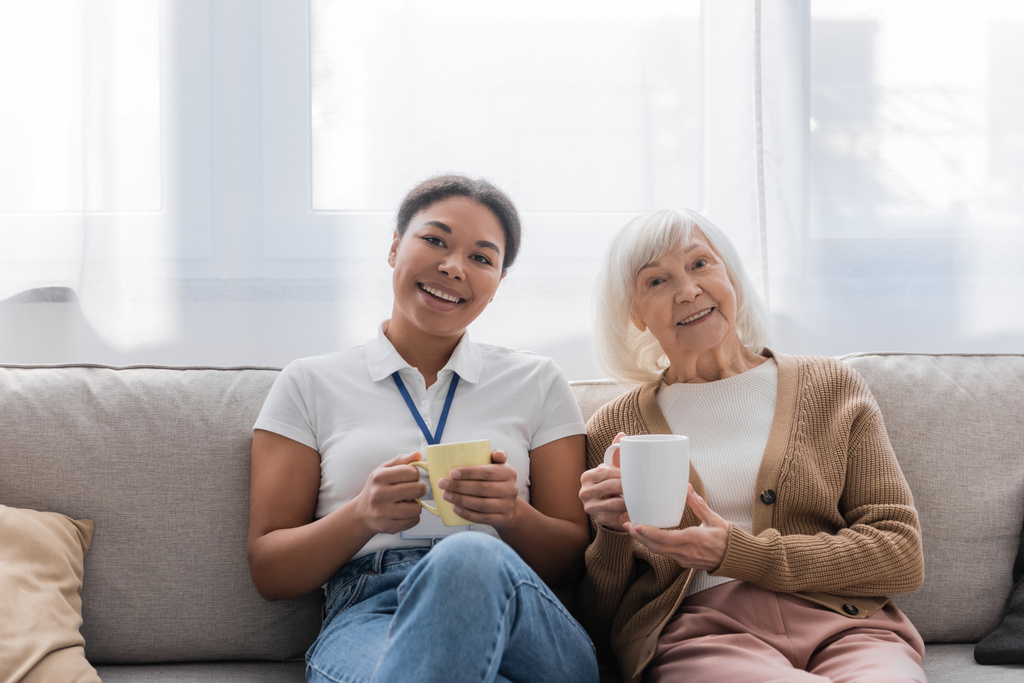 alegre trabajador social multirracial tomando el té con la mujer mayor en la sala de estar  - Foto, Imagen