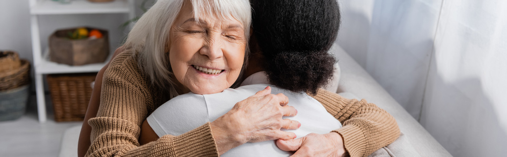 gelukkig senior vrouw met gesloten ogen knuffelen multiraciale maatschappelijk werker in woonkamer, banner  - Foto, afbeelding