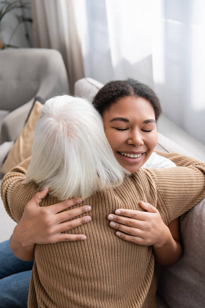fröhliche multirassische Sozialarbeiterin mit geschlossenen Augen, die sich mit einer Seniorin im Wohnzimmer umarmt  - Foto, Bild