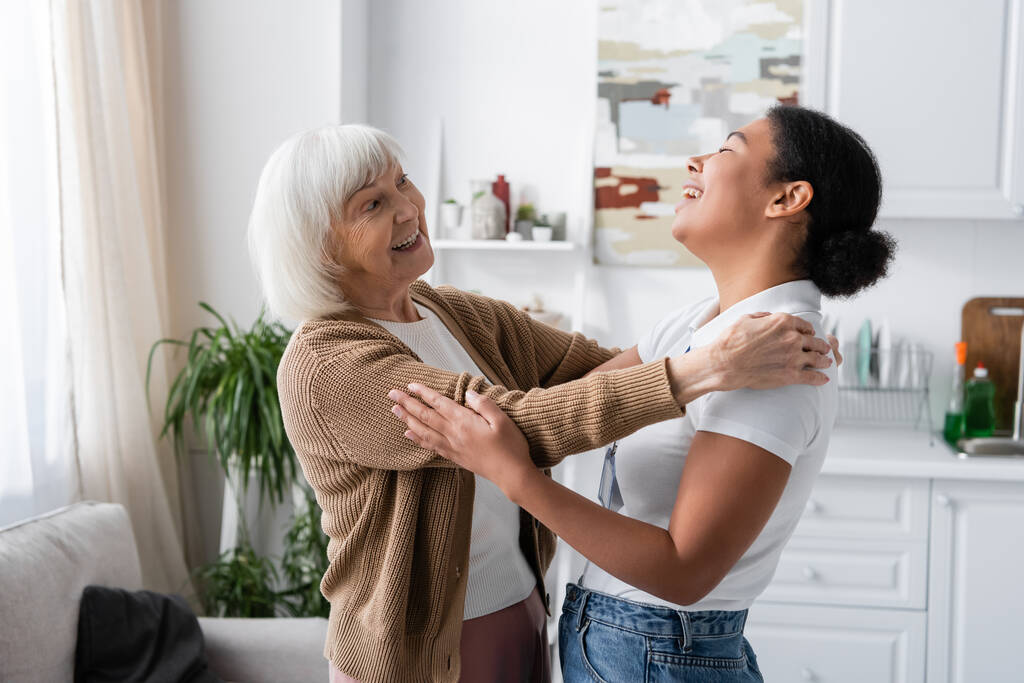 pozytywny wielorasowy pracownik socjalny i starsza kobieta śmieją się i przytulają w salonie  - Zdjęcie, obraz