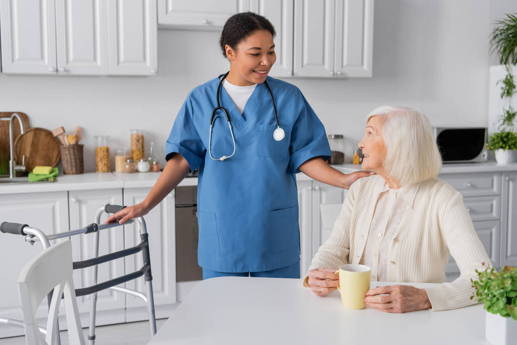 vidám nővér sztetoszkóppal nézi boldog idős nő ül egy csésze tea a konyhában  - Fotó, kép