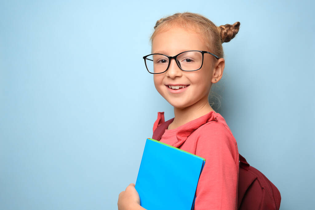 Мила дівчинка в окулярах, рюкзаку та підручнику на світло-блакитному тлі. Простір для тексту
 - Фото, зображення